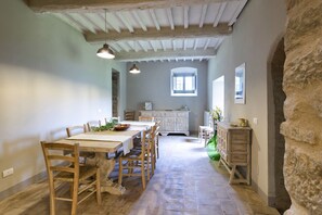 A rustic dining area with a wooden table and chairs, a stone wall, and a view of a kitchen in the background. - Villa Elda (Toscana)
