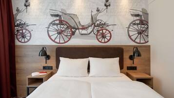 Une chambre d’hôtel avec un grand lit, deux tables de chevet, une petite table d’appoint, une fresque murale représentant une calèche tirée par des chevaux et un rideau rouge.