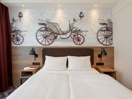 Una habitación de hotel con una cama grande, dos mesitas de noche, una mesita auxiliar y un mural en la pared con un carruaje tirado por caballos, además de una cortina roja.