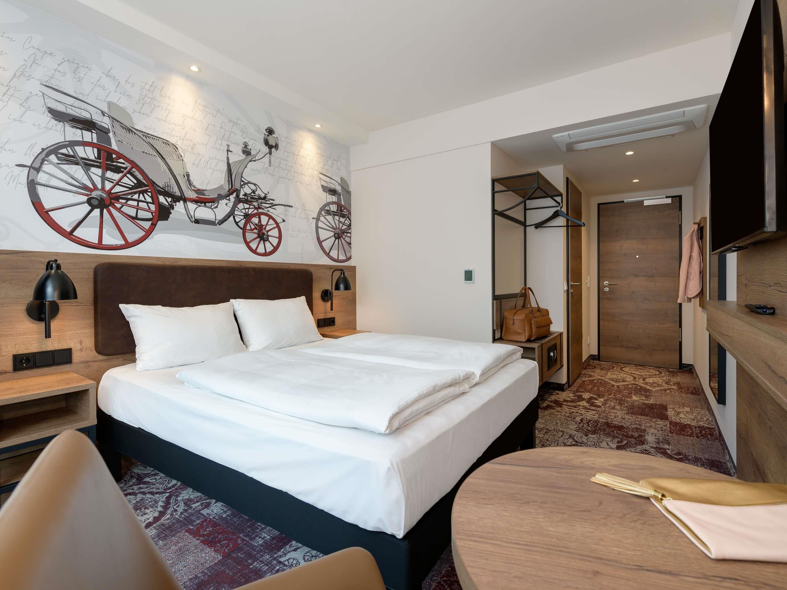 Une chambre d’hôtel comprenant un lit, des tables de chevet, un bureau, une chaise, une table ronde avec une bouteille, un porte-manteau, une armoire et un téléviseur.