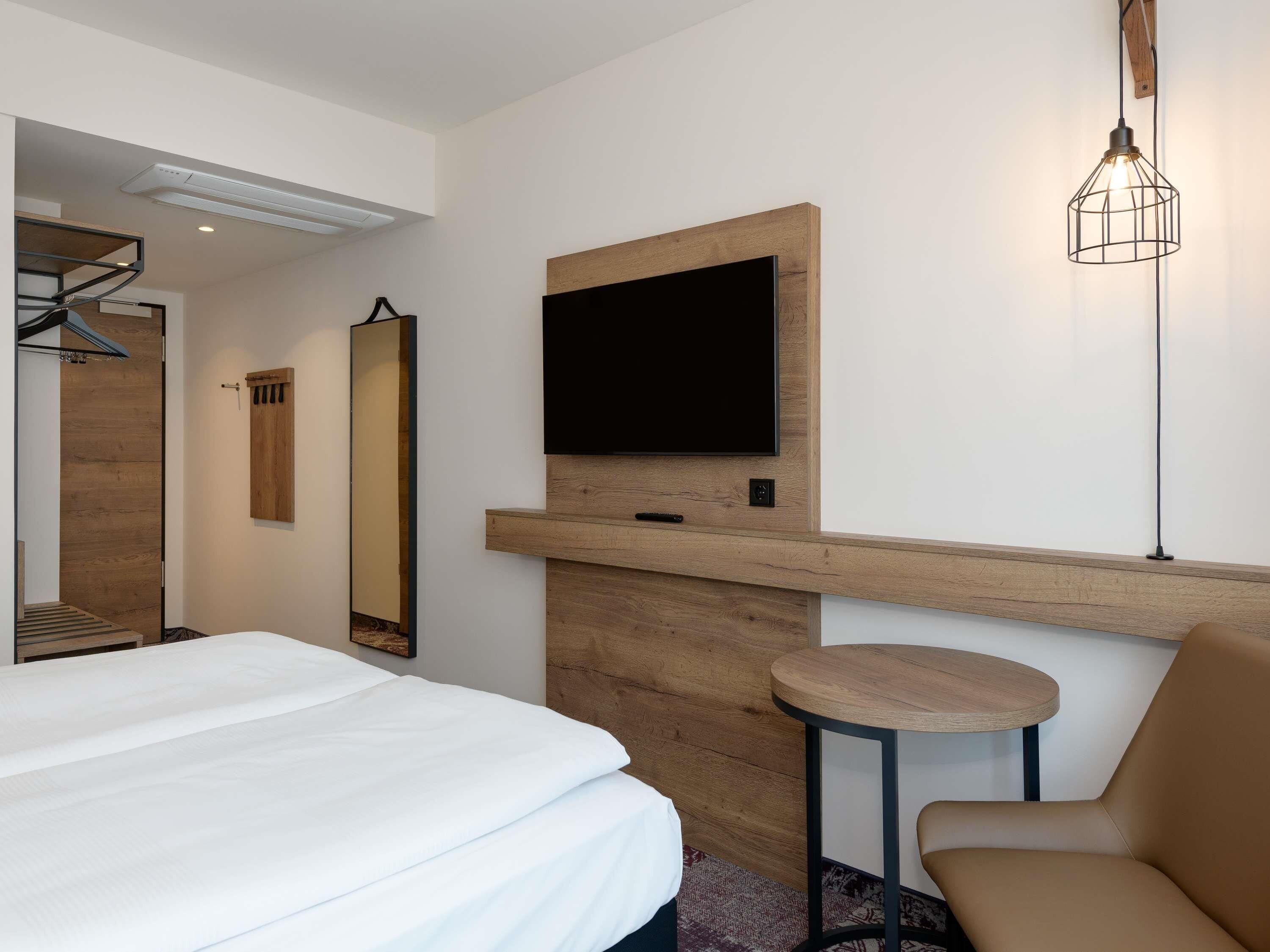 A modern hotel room with a bed, a mounted TV, a wooden accent wall, a mirror, and a small round table with a chair.