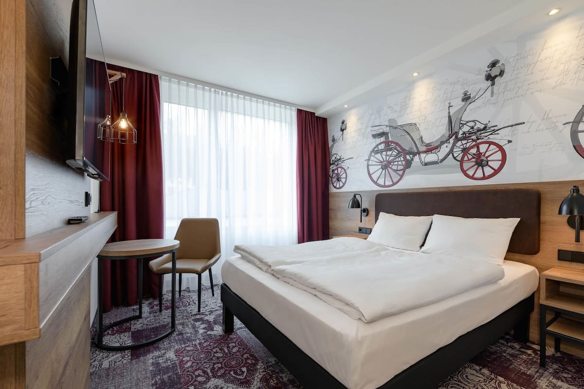 A hotel room with a bed, a desk, a chair, a TV, and a wall with bicycle artwork.