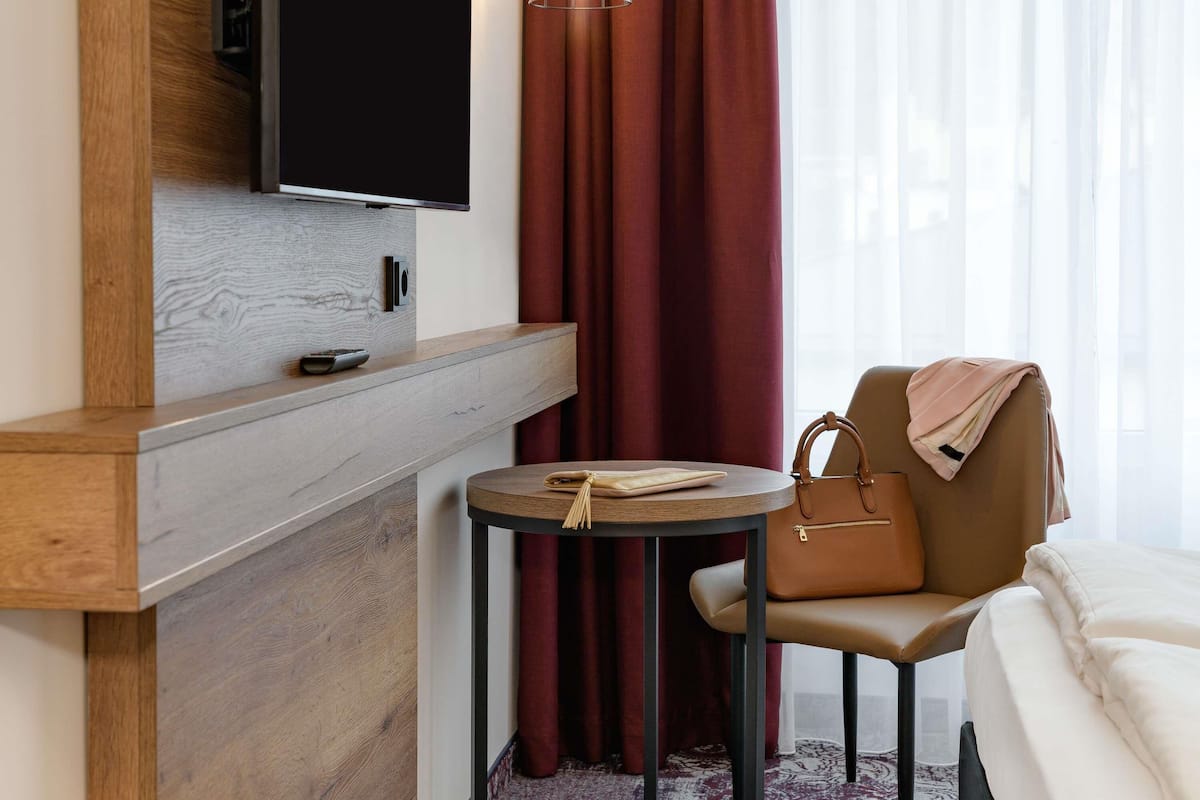 A hotel room with a bed, a TV mounted on a wooden panel, a small round table with a handbag, and a red curtain.