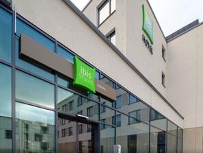 Un edificio moderno con fachada de vidrio, un letrero verde que dice 'ibis Styles COBURG' y un anuncio que indica 'SMILE Welcome en ibis Styles'.