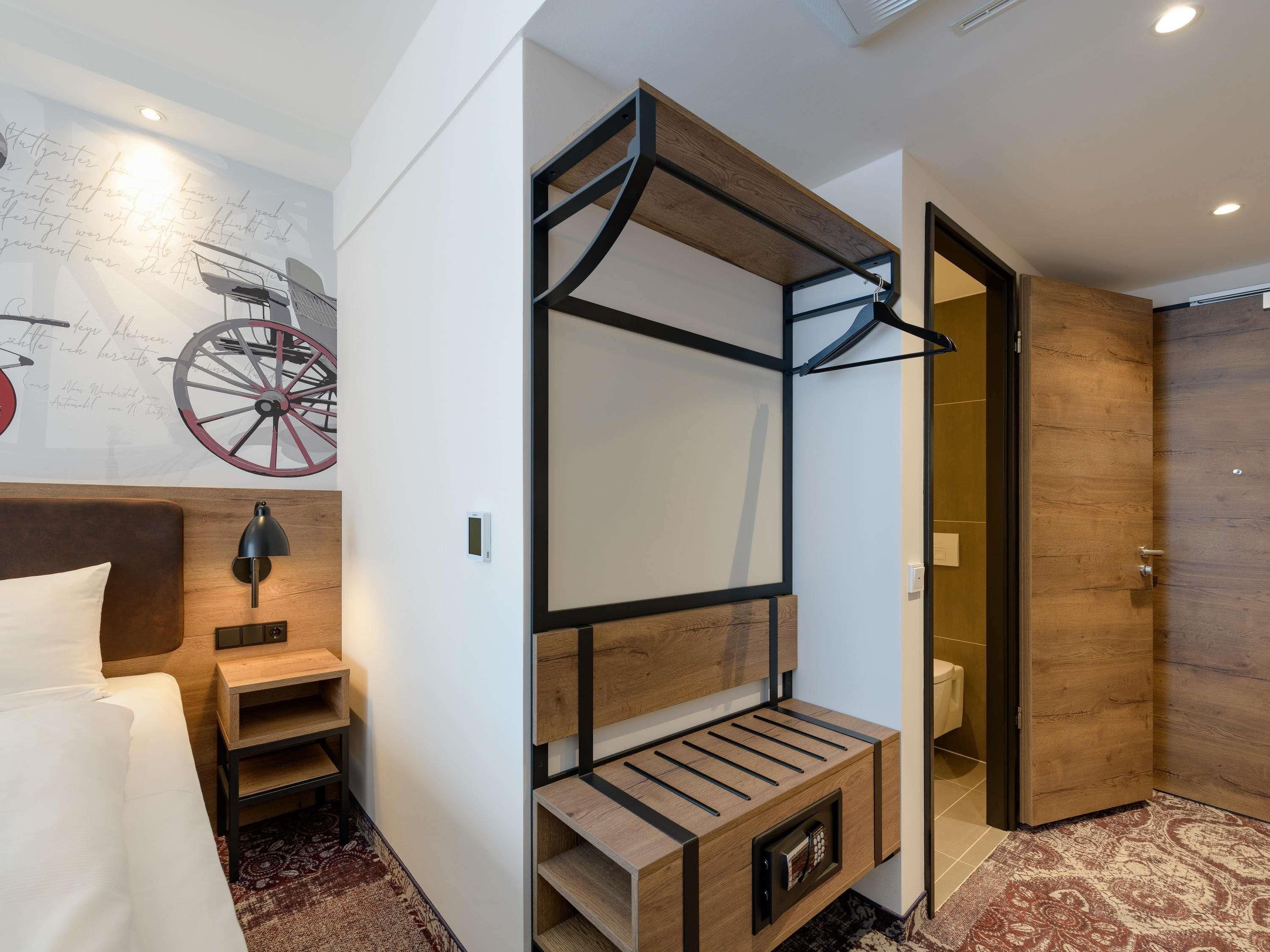 A modern hotel room with a bed, a wooden bench, a wall-mounted TV, and a door leading to another room.