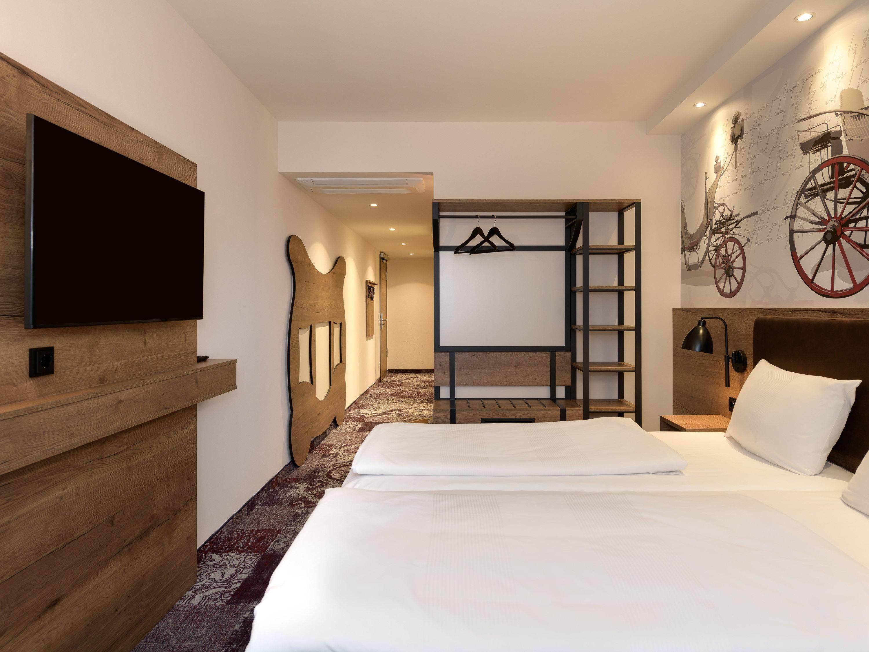 Une chambre d’hôtel avec un grand lit, un téléviseur à écran plat fixé au mur, une tête de lit en bois, une armoire avec un miroir et une étagère murale sur laquelle sont posés un vélo et une roue rouge.