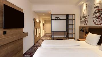 Une chambre d’hôtel avec un grand lit, un téléviseur à écran plat fixé au mur, une tête de lit en bois, une armoire avec un miroir et une étagère murale sur laquelle sont posés un vélo et une roue rouge.