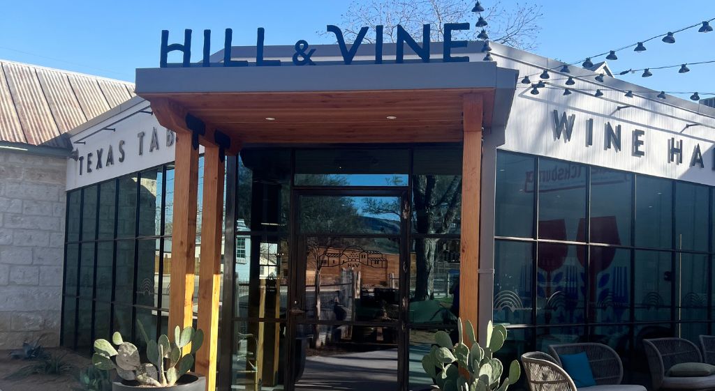 El exterior de Hill & Vine Wine House, con entrada despejada, zona de estar al aire libre y un letrero que indica que es un lugar para ir, comer y divertirse.