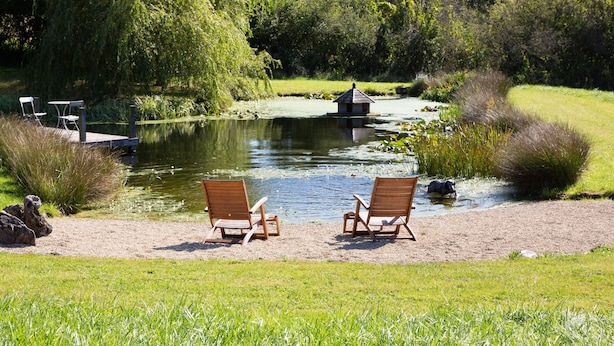 Een rustgevende oever met twee houten stoelen gericht op het water, een klein houten gebouw aan de waterkant, en weelderige begroeiing op de achtergrond.