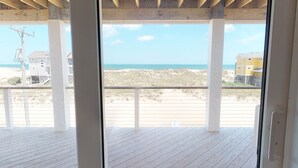 On peut admirer la plage depuis une terrasse en bois dotée d’une porte vitrée, avec vue sur la plage et un bâtiment situé au loin.