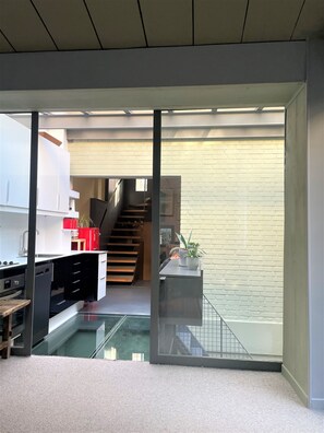 A modern kitchen with a glass partition, a staircase leading to a loft, and a brick wall.