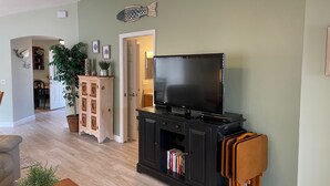 A living room with a flat-screen TV on a black stand, a wooden cabinet with books, a potted plant, and a dining area visible in the background.