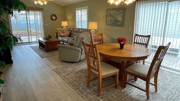 A spacious living room with a sofa, a dining table with chairs, a ceiling fan, and a large window with blinds.