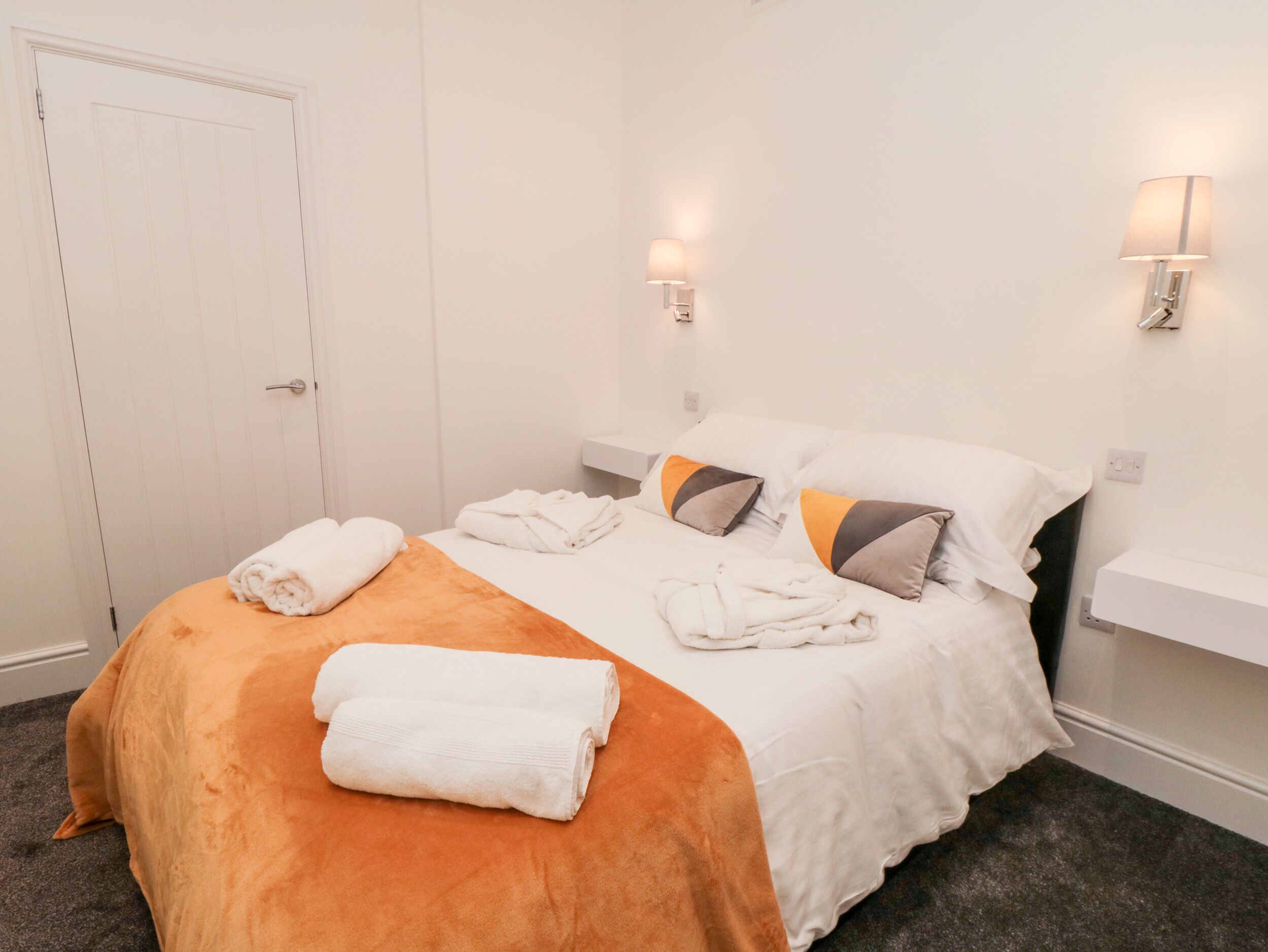 A neatly made bed with white linens and an orange bedspread, two wall-mounted lights, and a white door.