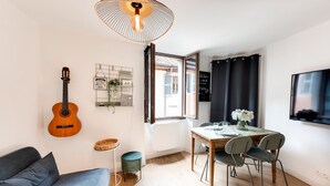 A modern living room with a guitar, a dining table with chairs, a sofa, a small side table, and a large window with curtains.