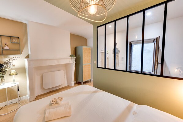 Une chambre d’hôtel avec un lit, une petite table avec une lampe, une étagère fixée au mur et une fenêtre avec des stores.