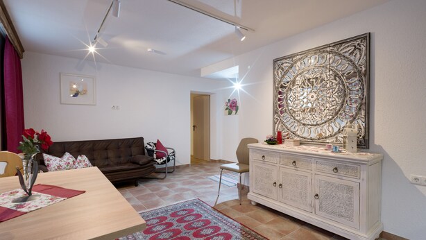 A modern living room with a brown leather sofa, a dining table with a vase of red flowers, a patterned rug, and a decorative wall piece. - Urlaub in den Bergen (Uderns)
