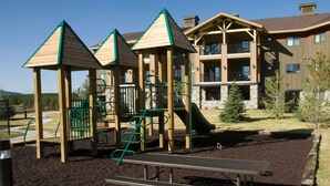 A playground with climbing structures and a slide, surrounded by a landscaped area with a building in the background.