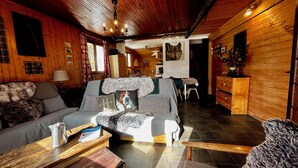 A cozy cabin interior with a wooden interior, a sofa with a fur throw, a dining table, and a kitchen area in the background.