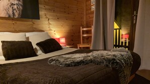 A wooden cabin room with a bed, pillows, a blanket, a nightstand with a lamp, and a wooden chair.