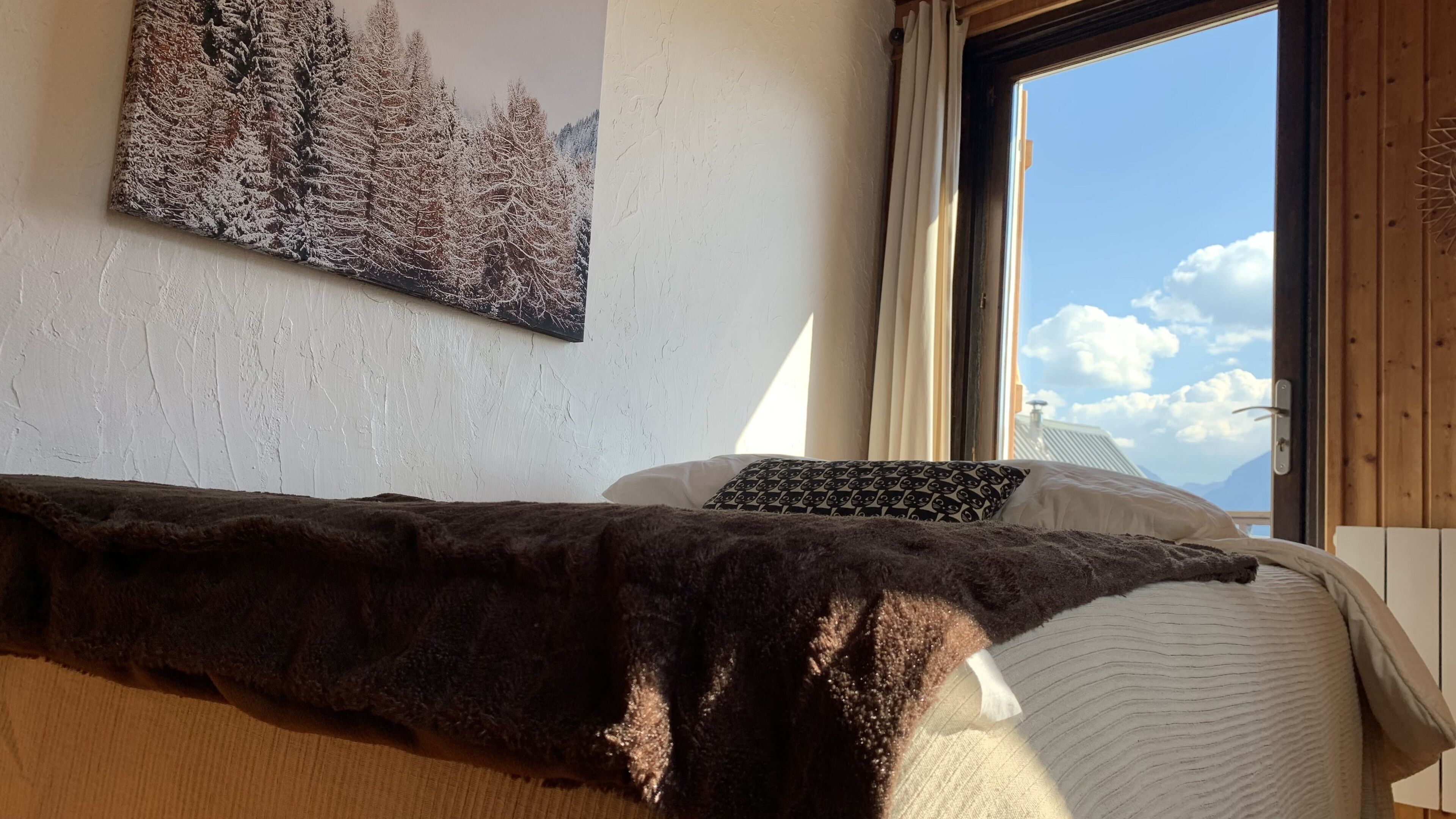 Ett sovrum med en säng, ett fönster med utsikt över himlen och molnen, och en tavla med träd på väggen.