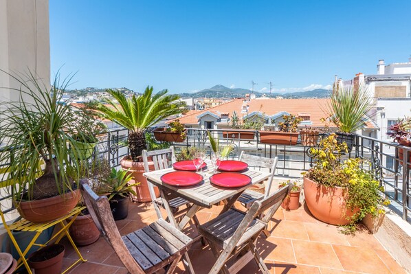 A terrace with a table set for four, surrounded by potted plants, overlooking a cityscape with buildings and mountains. - Happyfew La terrasse de Rossini (Nice)
