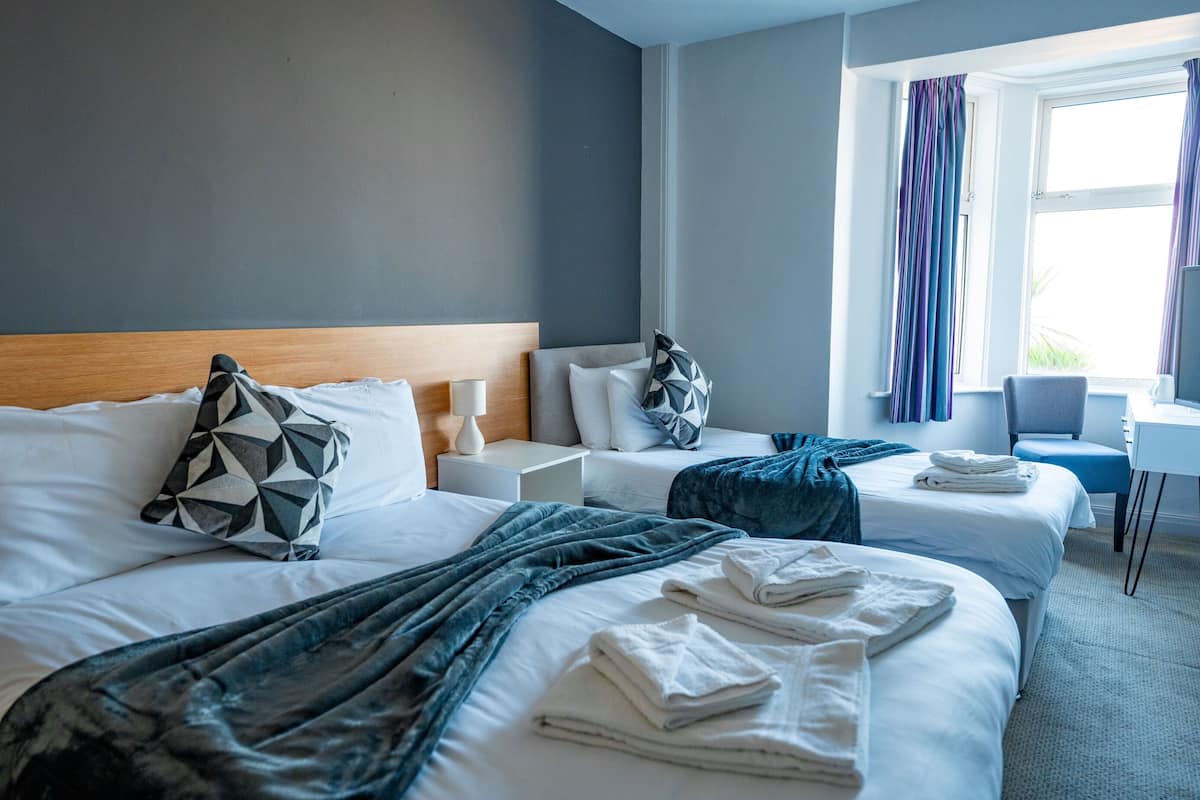 Habitación de hotel con dos camas, cabecero de madera, ropa blanca, manta azul y una ventana con cortinas.