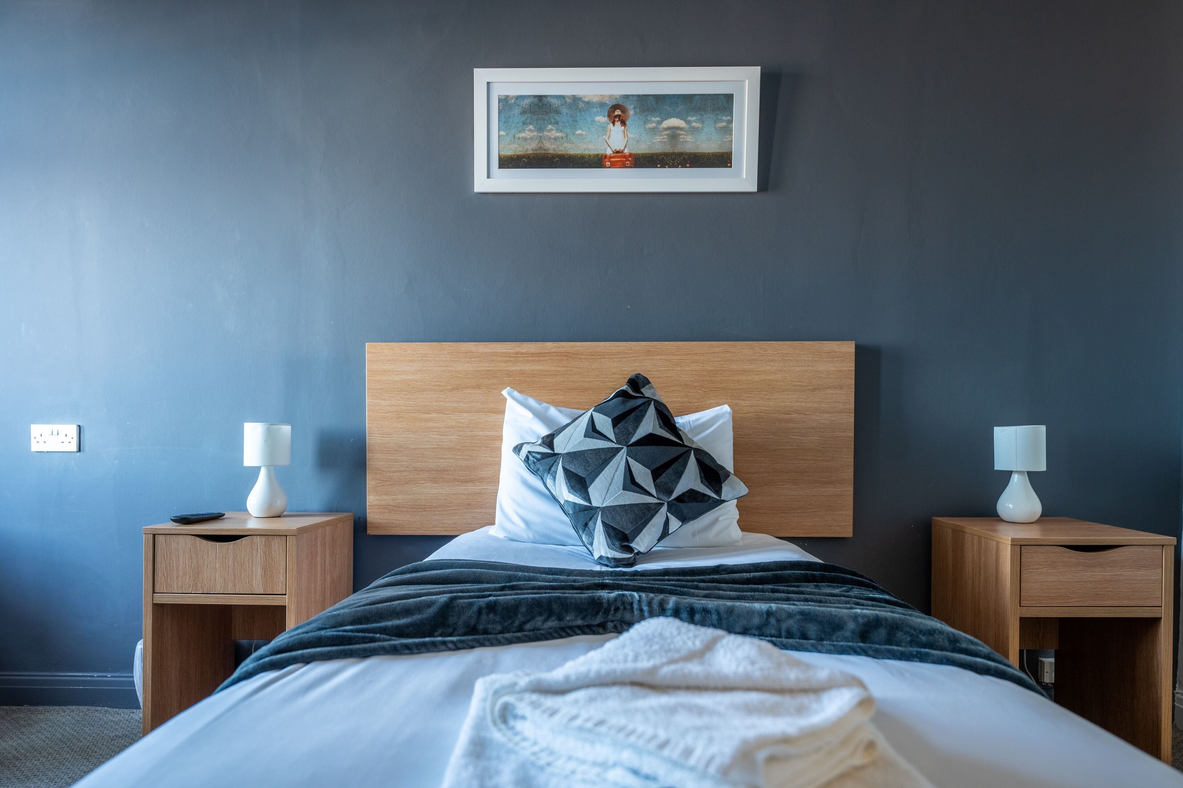 A modern hotel room with a large bed, two bedside tables, a nightstand, a framed picture on the wall, and a dark blue wall.