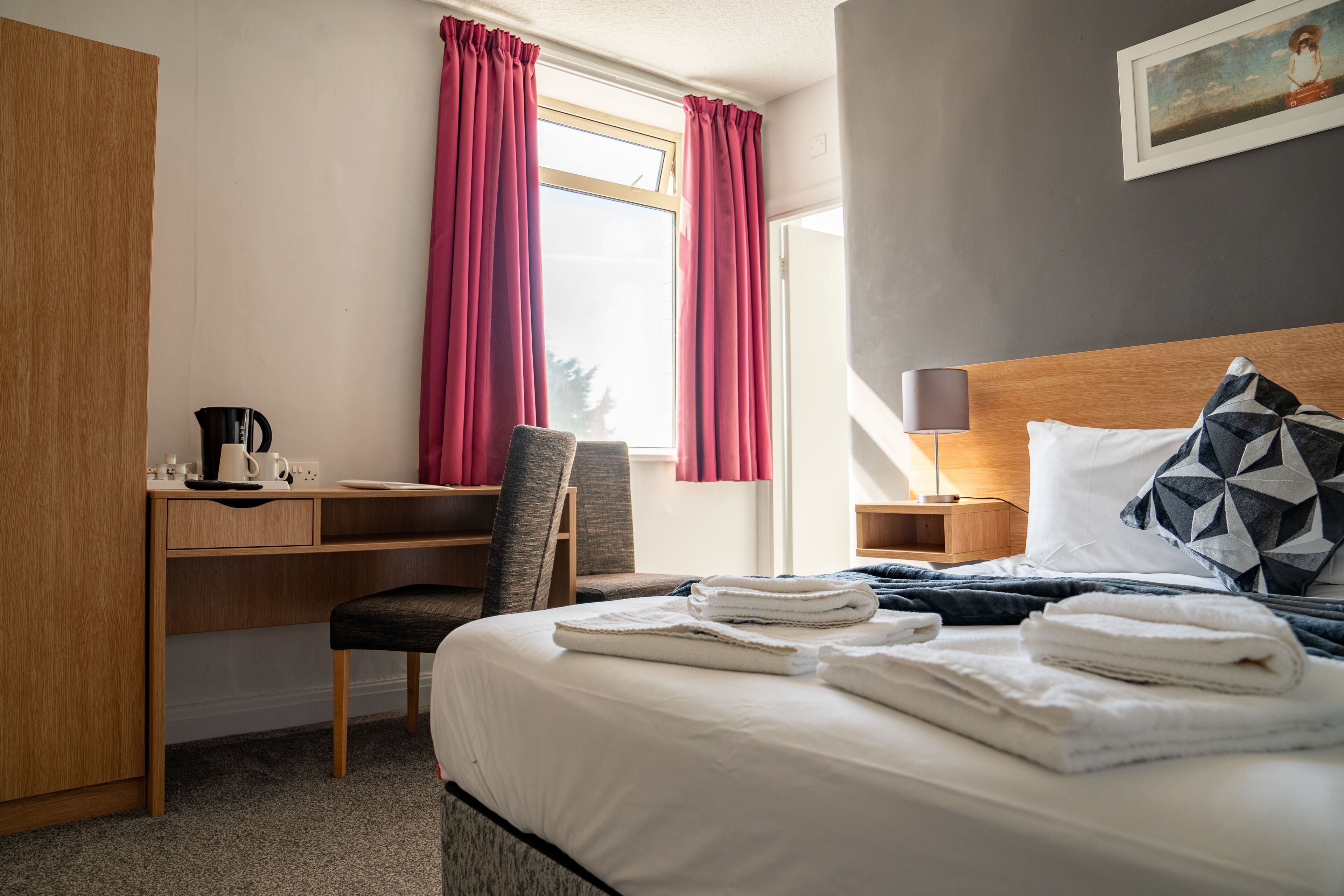 Una habitación de hotel con cama, escritorio, silla, ventana con cortinas, mesita de noche y lámpara.
