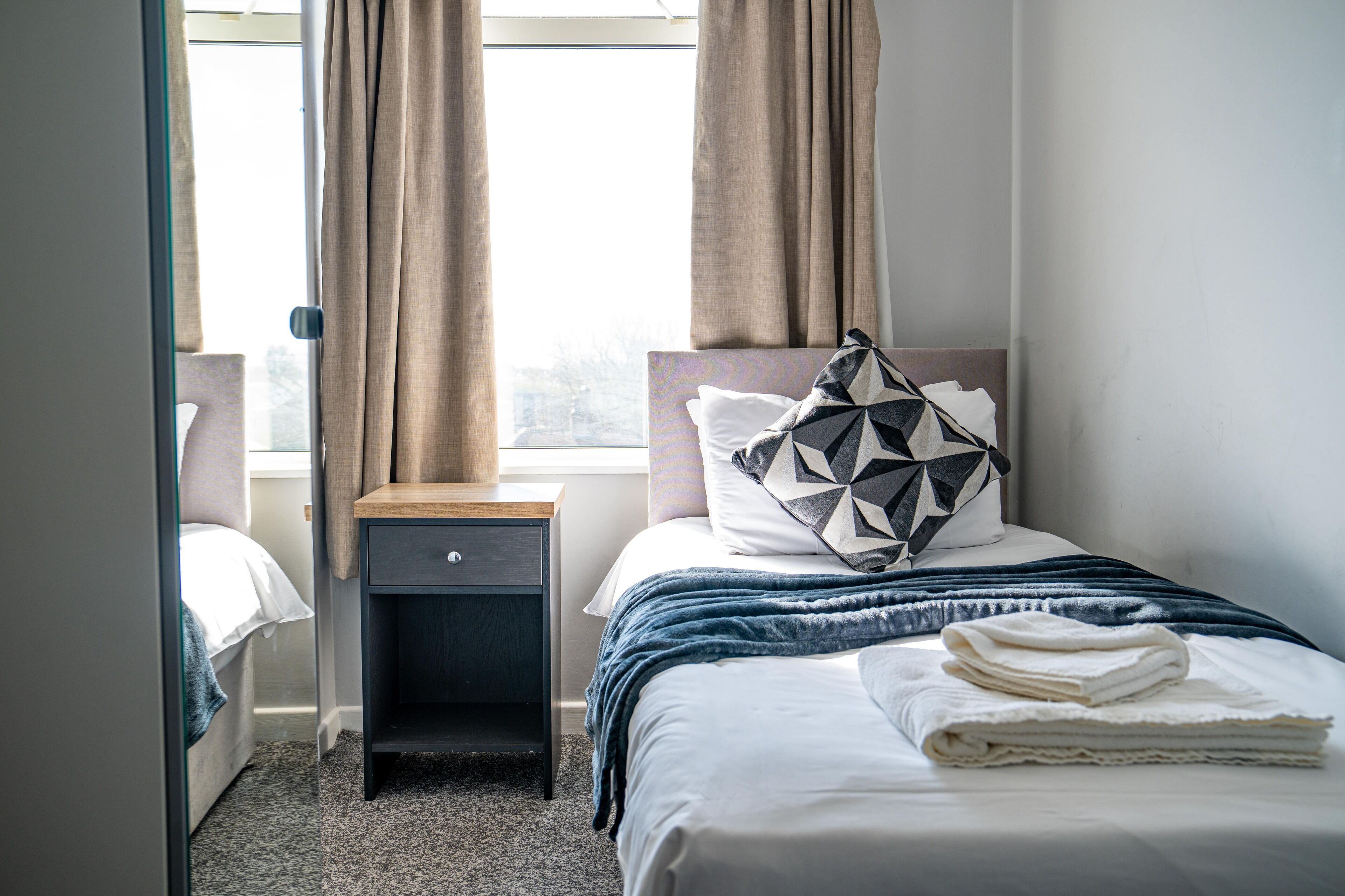A neatly made bed with a geometric pillow, a bedside table, and a window with curtains.