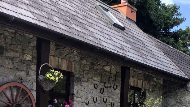 Ein Steinhaus mit einem geneigten Dach, einem hölzernen Wasserrad und verschiedenen Blumentöpfen und Blumen.