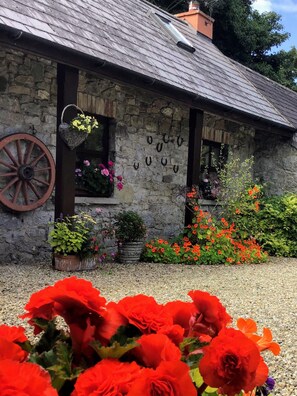 Un bâtiment en pierre avec un toit en pente, une roue en bois, ainsi qu’une variété de plantes et de fleurs en pot.