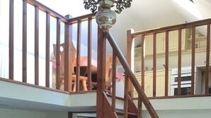 Eine mehrstufige Holztreppe verbindet zwei Etagen. Im Vordergrund steht ein hölzernes Bett, daneben ein Holzschrank, ein Spiegel und ein Fenster mit Blick auf die obere Etage.