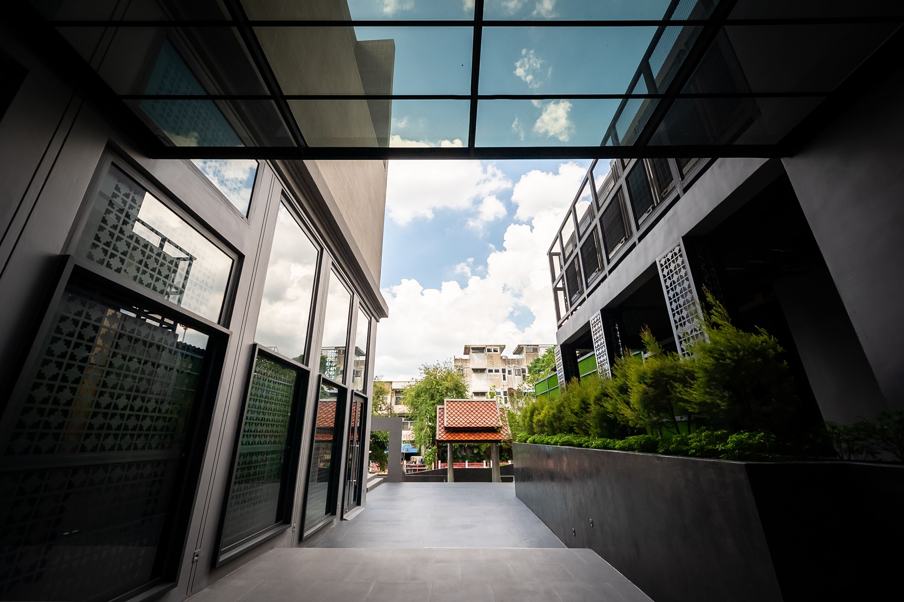 Een modern gebouw met grote ramen en een glazen dak, uitlopend in een binnenplaats met een constructie met een rood dak en beplanting.
