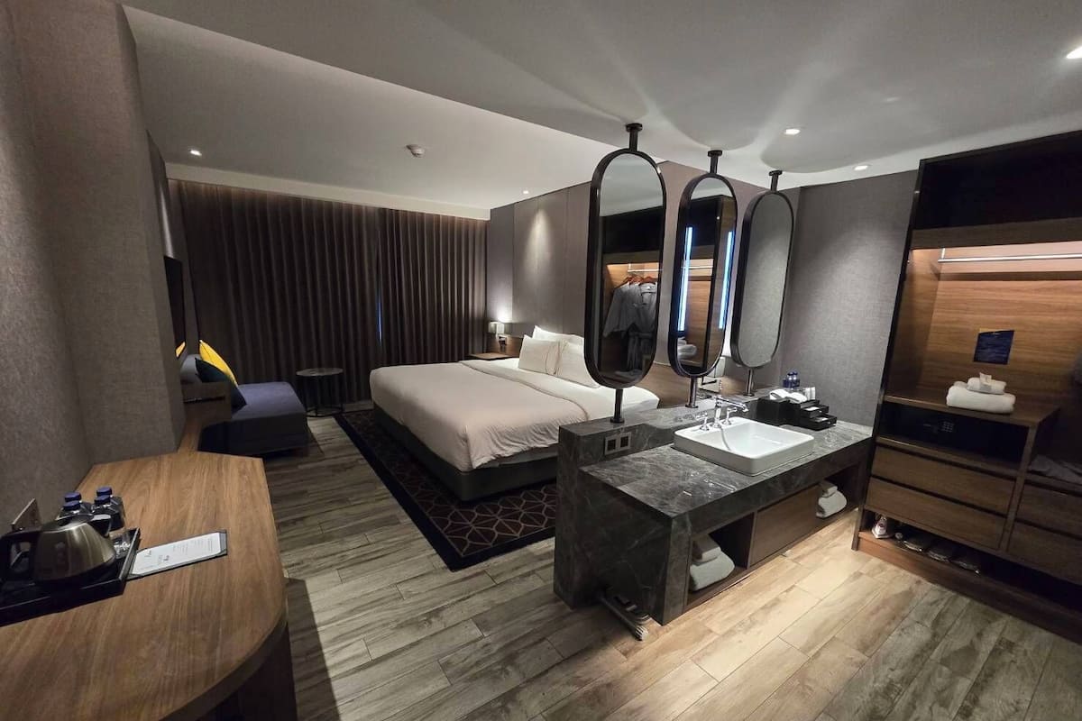 A hotel room with a bed, a vanity with a mirror, a sink, a wooden wardrobe, and a small table with a kettle.