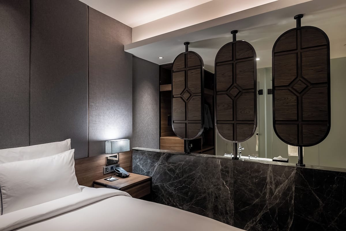 A hotel room with a bed, a nightstand with a lamp, a marble countertop, and a large mirror with wooden panels.