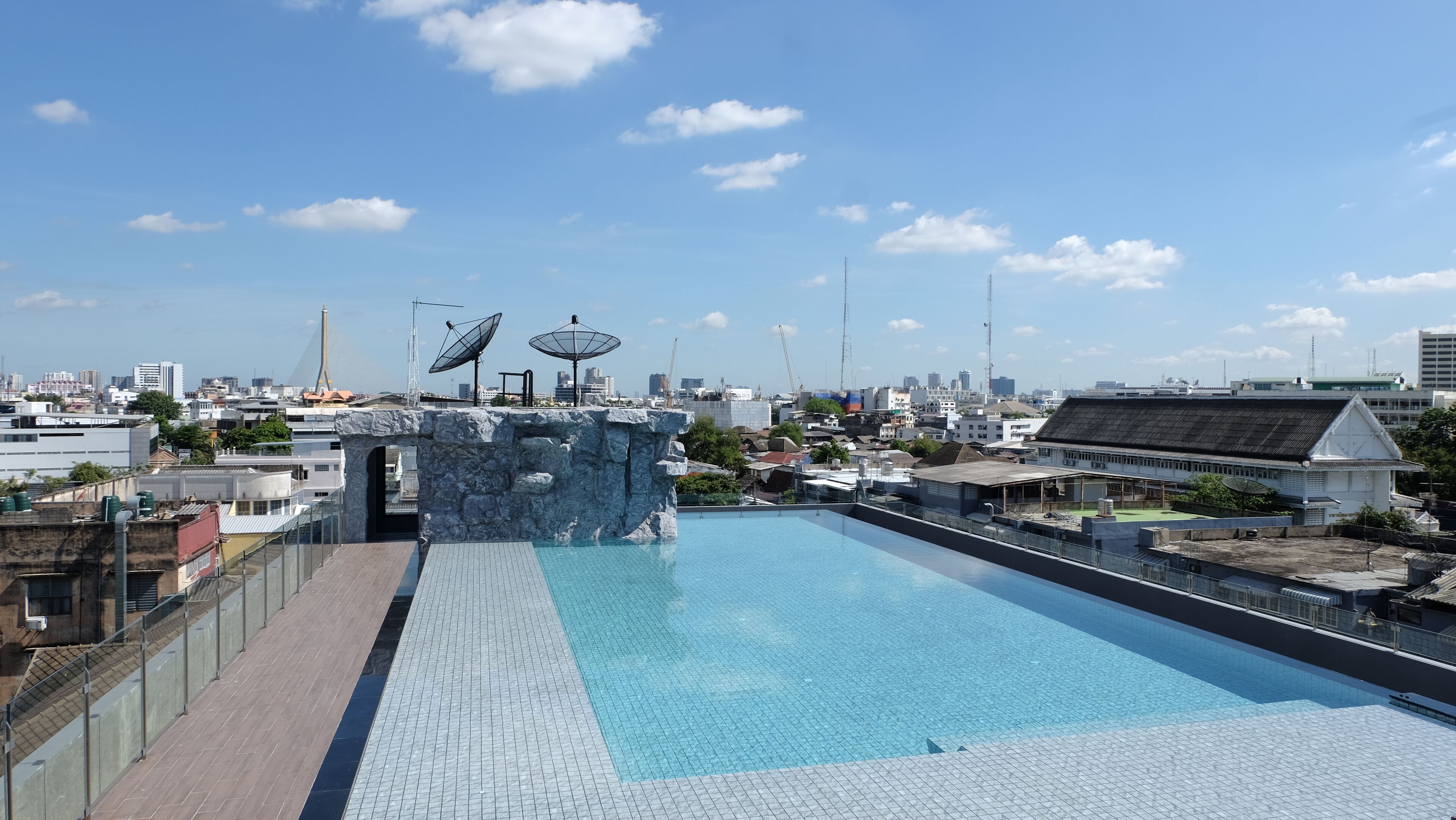 Une piscine sur le toit, agrémentée d’une arche en pierre et d’antennes paraboliques, offrant une vue sur un paysage urbain composé de différents bâtiments et d’un pont situé au loin.