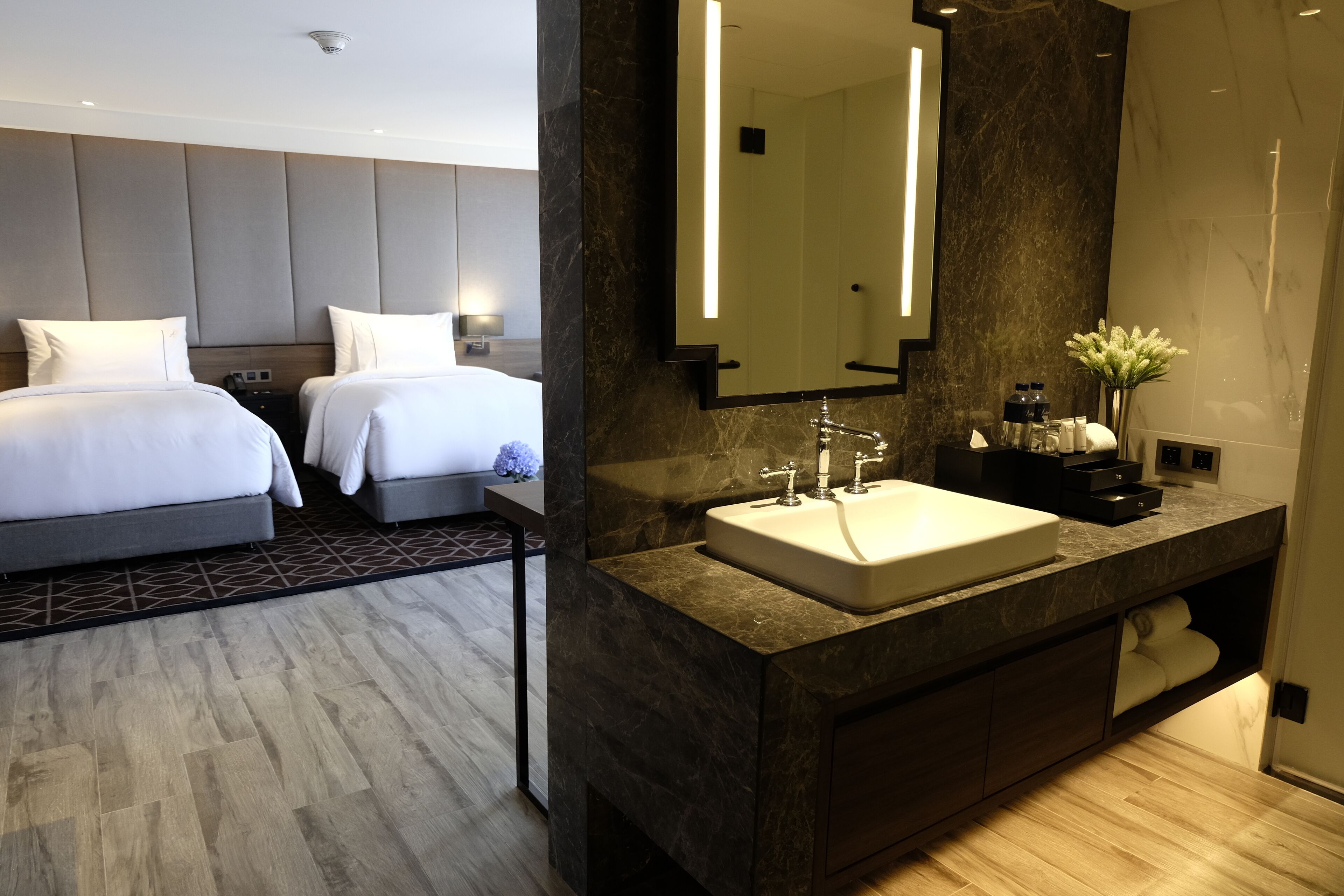 A modern hotel room with two beds, a marble bathroom vanity, and a large mirror.