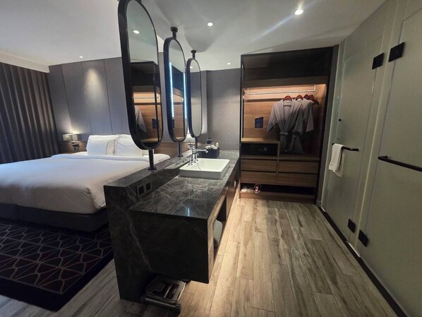 A hotel room with a bed, a bathroom with a sink and mirror, and a walk-in closet with hanging clothes. - Ayathorn Bangkok (Bangkok)