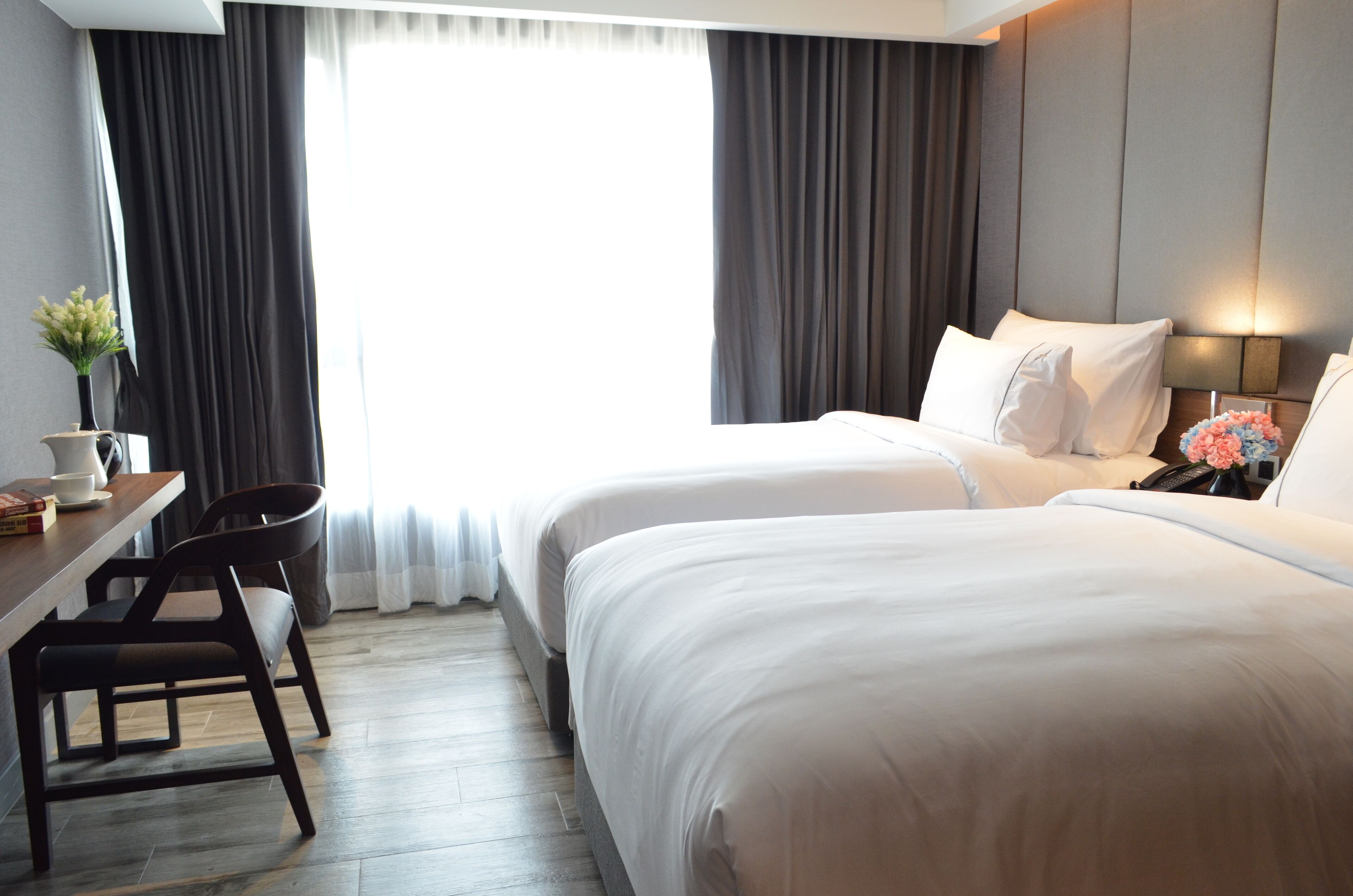 A hotel room with two beds, a desk with chairs, a small table with a vase, and a window with curtains.