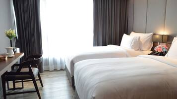 A hotel room with two beds, a desk with chairs, a small table with a vase, and a window with curtains.