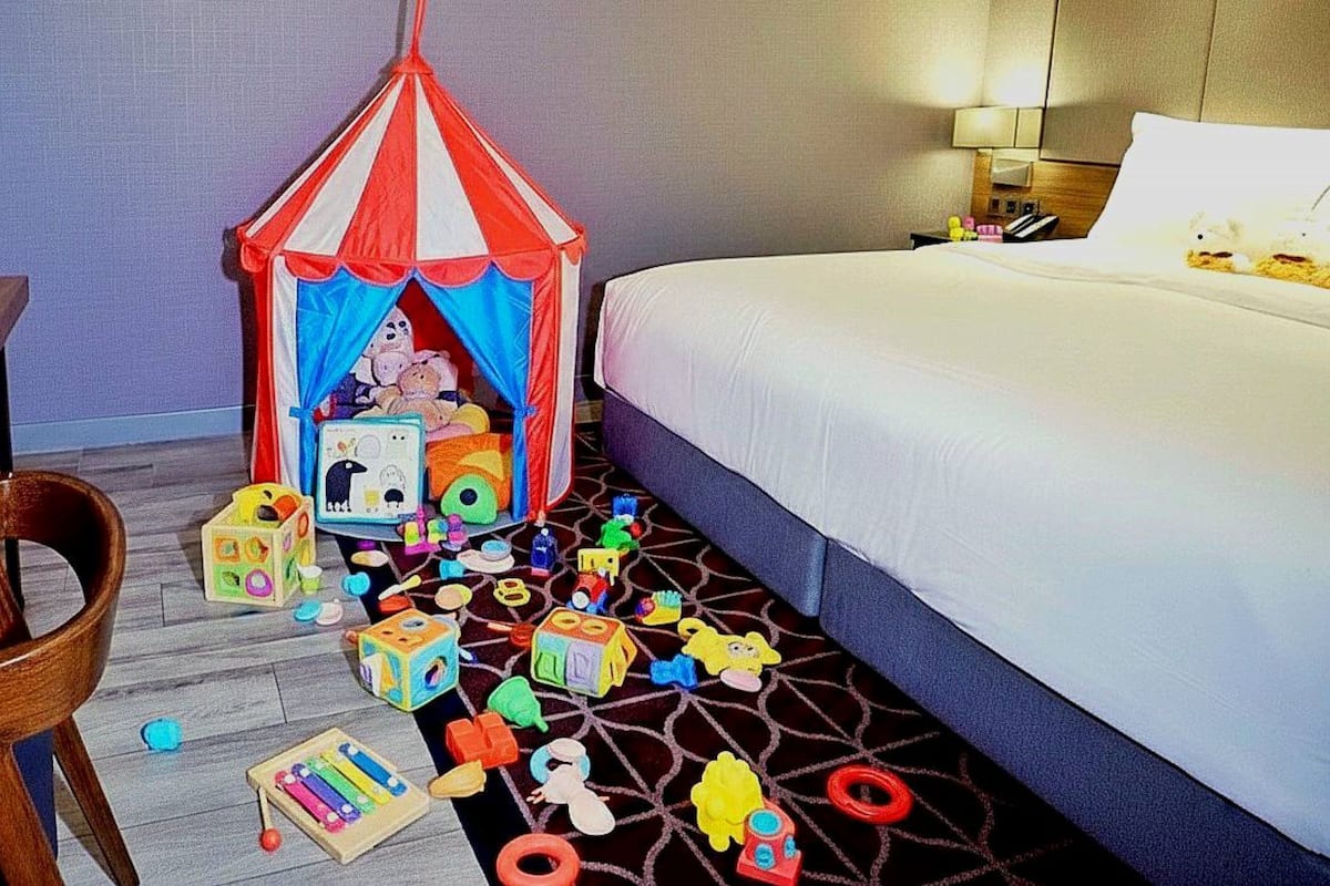 A hotel room with a bed, a small play tent, and scattered toys on the floor.