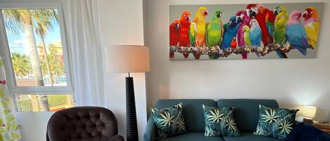 A living room with a sofa, a chair, a painting of colorful parrots, a lamp, and a view of palm trees through a window.