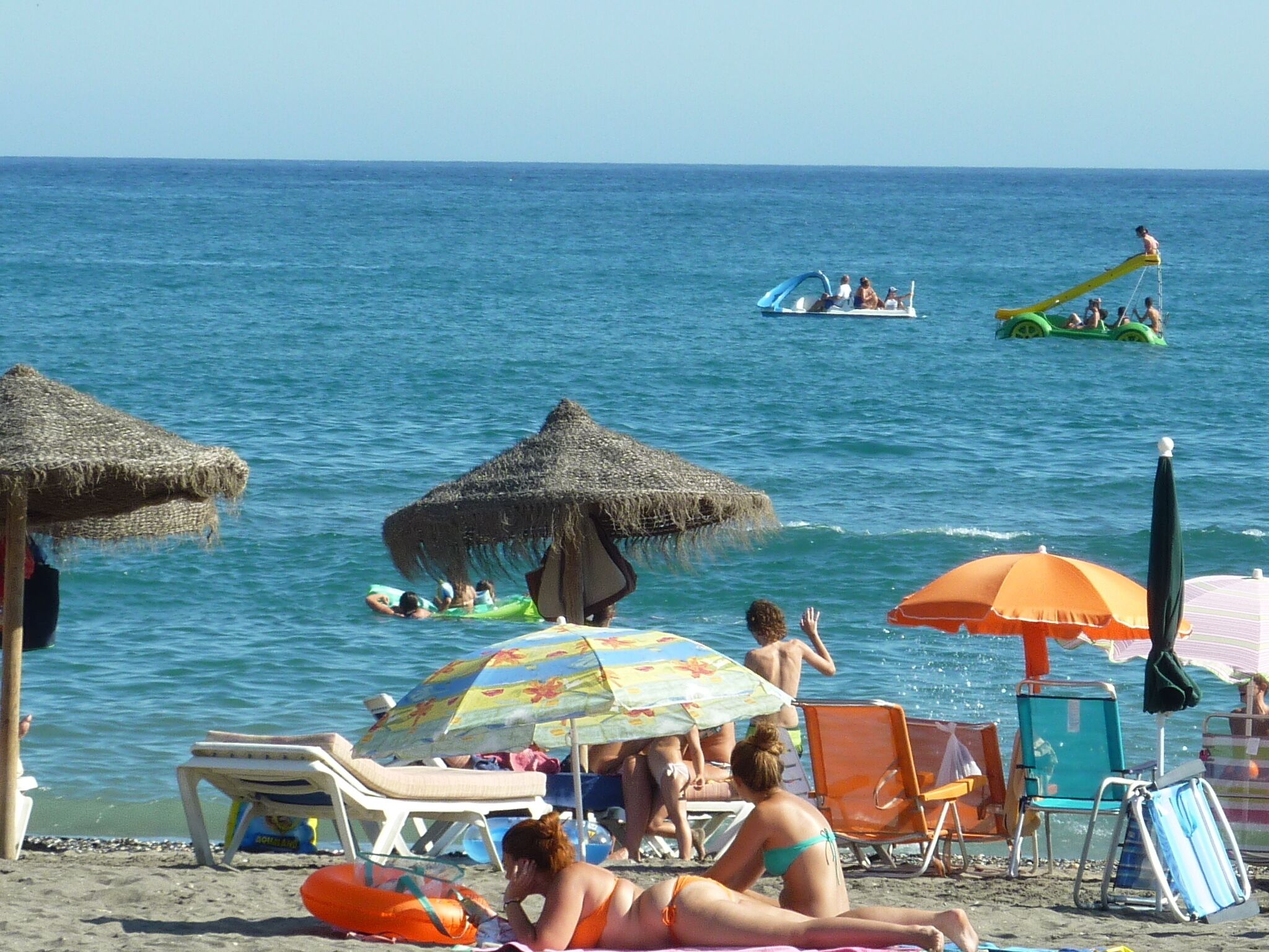 Une plage avec des parasols en chaume, des chaises longues et des gens qui se détendent au bord de l’eau.