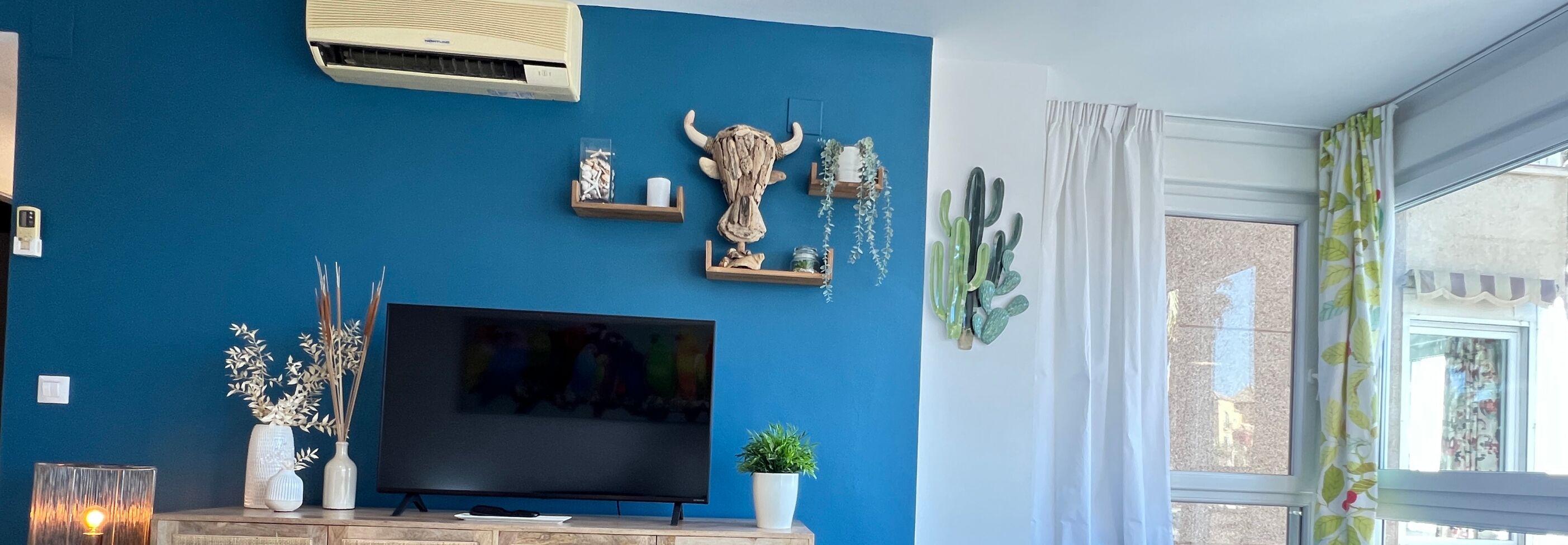 A living room with a blue wall, a mounted air conditioner, a flat-screen TV, decorative items including a bull head sculpture, and a window with curtains.