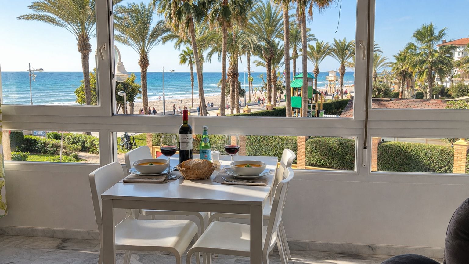 A dining table with two bowls of soup, a bottle of wine, and a basket of bread, overlooking a beach with palm trees and the ocean.