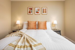 A neatly made bed with a white comforter and two orange pillows, flanked by two bedside tables with lamps, in a room with beige walls and three framed artworks above the bed.