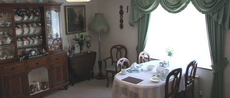 A dining room with a table set for four, surrounded by chairs. A wooden cabinet with glass doors displays various dishes. A chandelier hangs from the ceiling, and a window with green curtains is visible.