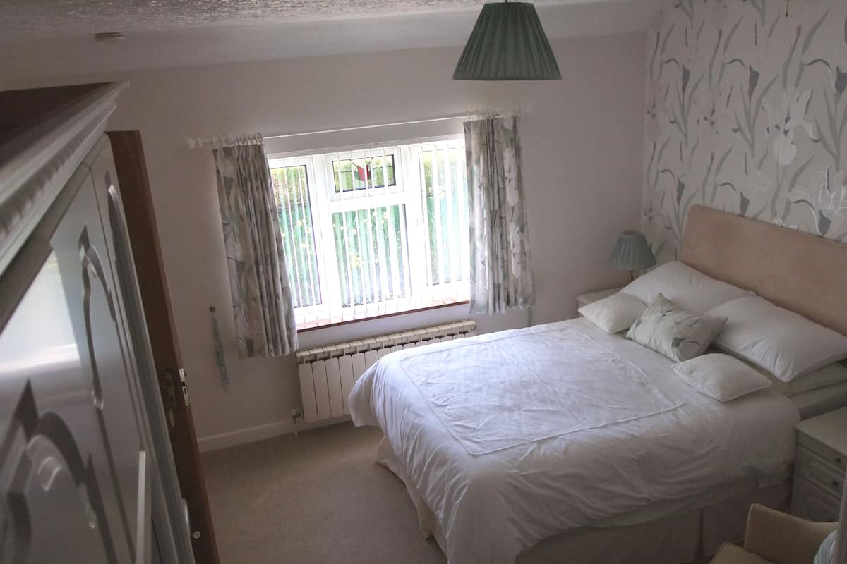 A bedroom with a large bed, a window with curtains, a radiator, and a nightstand with a lamp.