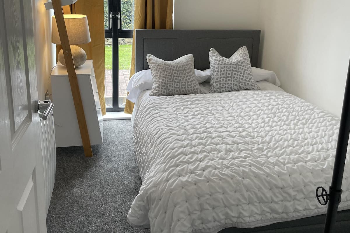 A bedroom with a bed, two pillows, a bedside table, a lamp, and a window with curtains.